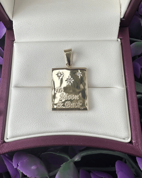 Rectangular gold pendant with engraved stars and an inscription 'To the moon & back', displayed in a jewellery box among purple flowers